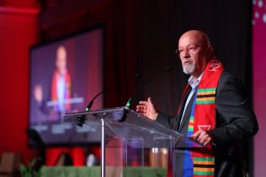 Christopher Lewis on stage making acceptance speech wearing a colourful shawl