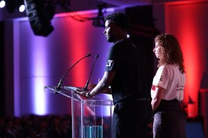 Young man and woman on stage, pictured from behind making a speech.